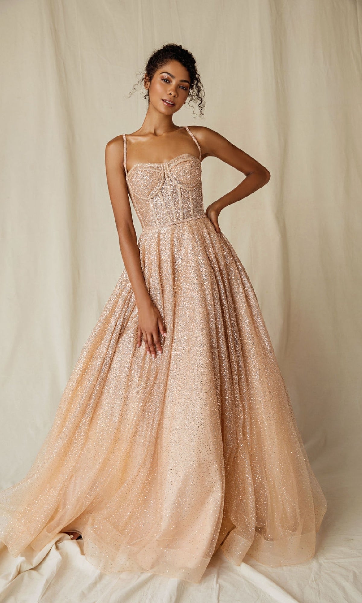 A woman poses before a beige backdrop in the Sheer-Corset Glitter Prom Ball Gown: Eureka 9900, featuring a sleeveless peach design with a sparkling corset-style bodice and full tulle skirt.